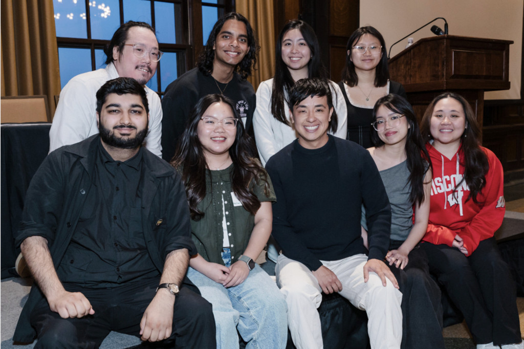 A group gathers around the stage for a photo. The first row sits on the stage with a row of attendees kneeling behind.